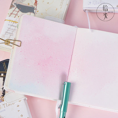 The River of Time Leather Journal with Magnetic Buckle, featuring a pastel watercolor interior and displayed with a green fountain pen on a pink background.