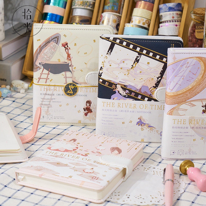 The River of Time Leather Journals with Magnetic Buckles displayed on a desk with various colorful covers featuring whimsical illustrations and surrounded by stationery items.