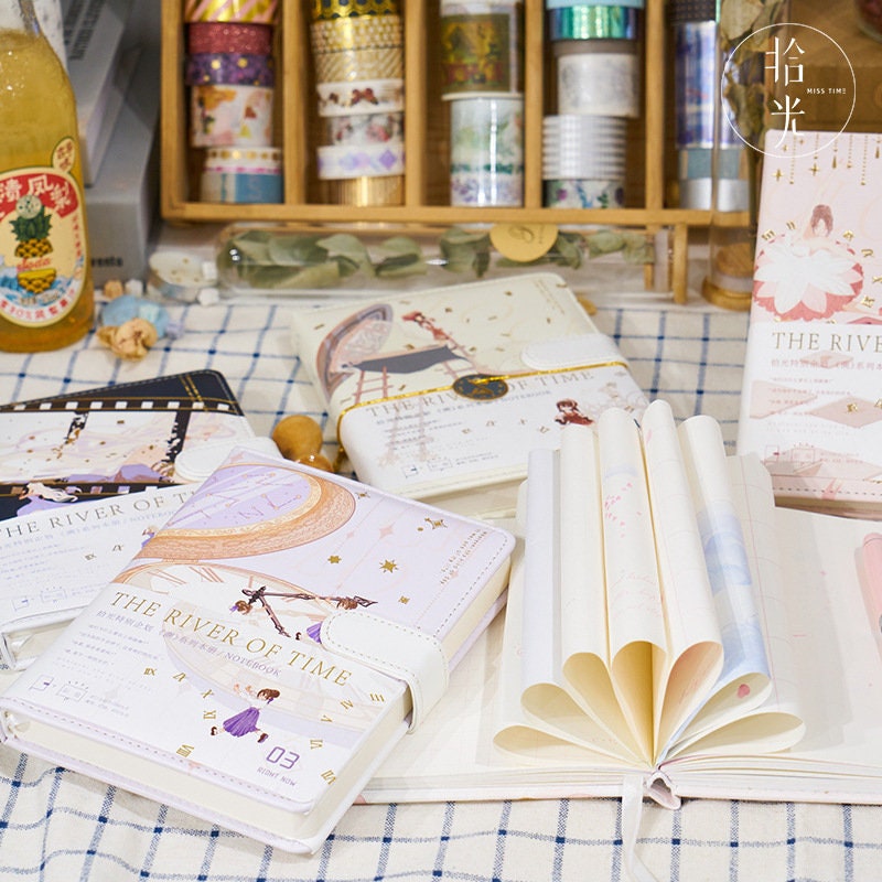 The River of Time Leather Journal with Magnetic Buckle in various designs displayed on a table with other stationery items and decorative washi tapes.