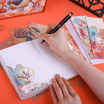 Person writing in a Blue Whale Notebook from the Blue Whale Notebook Set with Lighting Box, surrounded by colorful, artistic stationery on a bright orange surface.