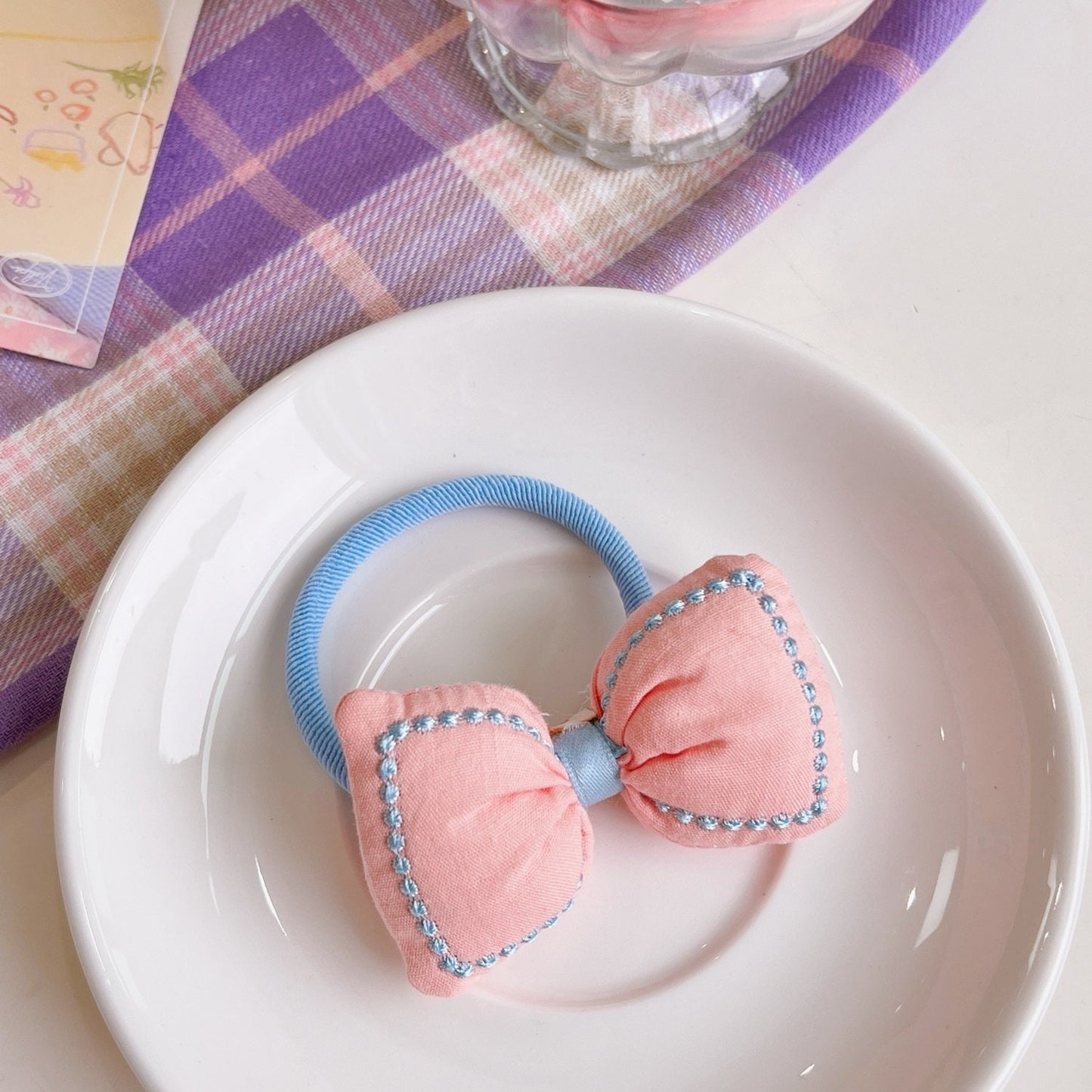 Kawaii Bowknot Hair Tie and Bracelet Combo in pastel pink with blue trim displayed on a white plate with checkered table cloth in the background.