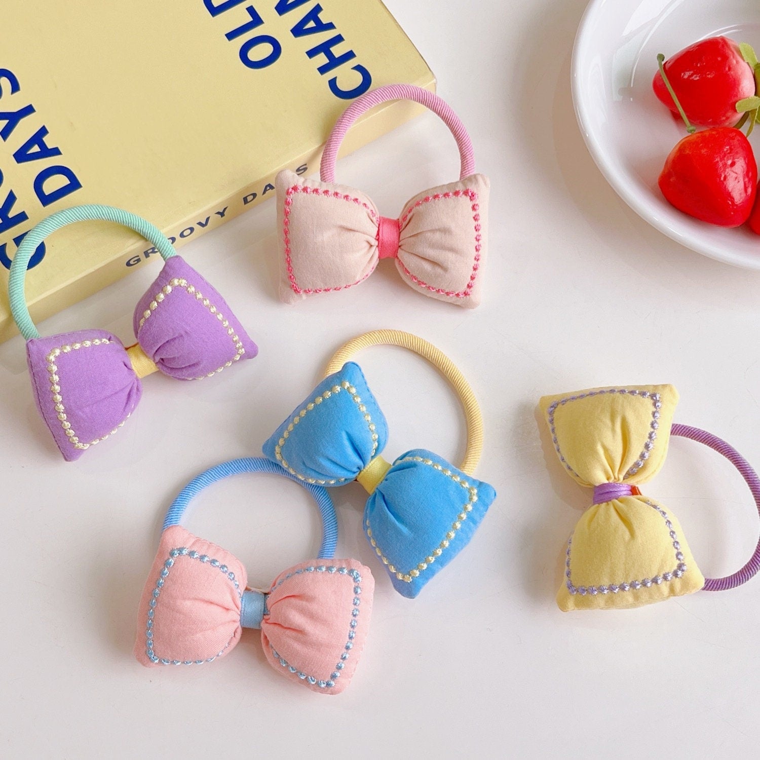 Kawaii Bowknot Hair Tie and Bracelet Combo in pastel colors including lavender, pink, blue, yellow, and peach, displayed on a desk with a book and faux cherries.