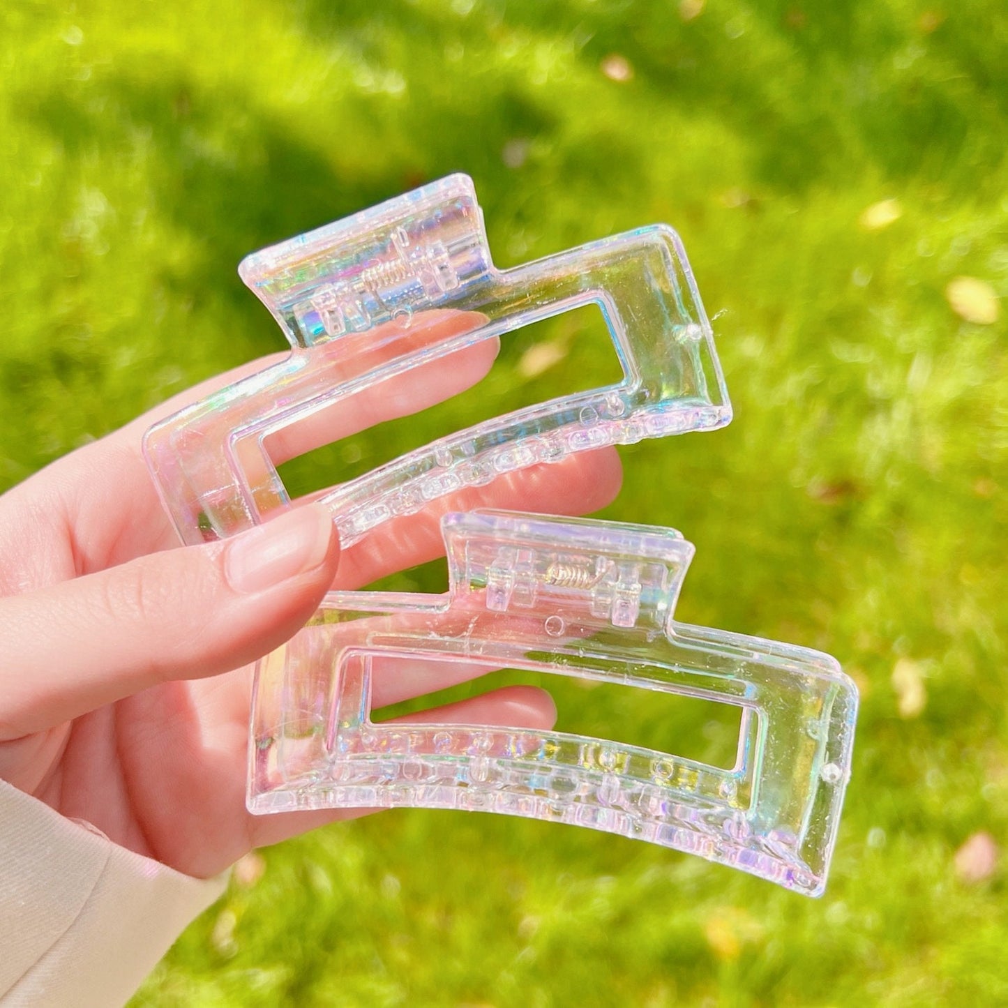 Transparent rainbow acrylic hair claw clip held by hand against green grass background.