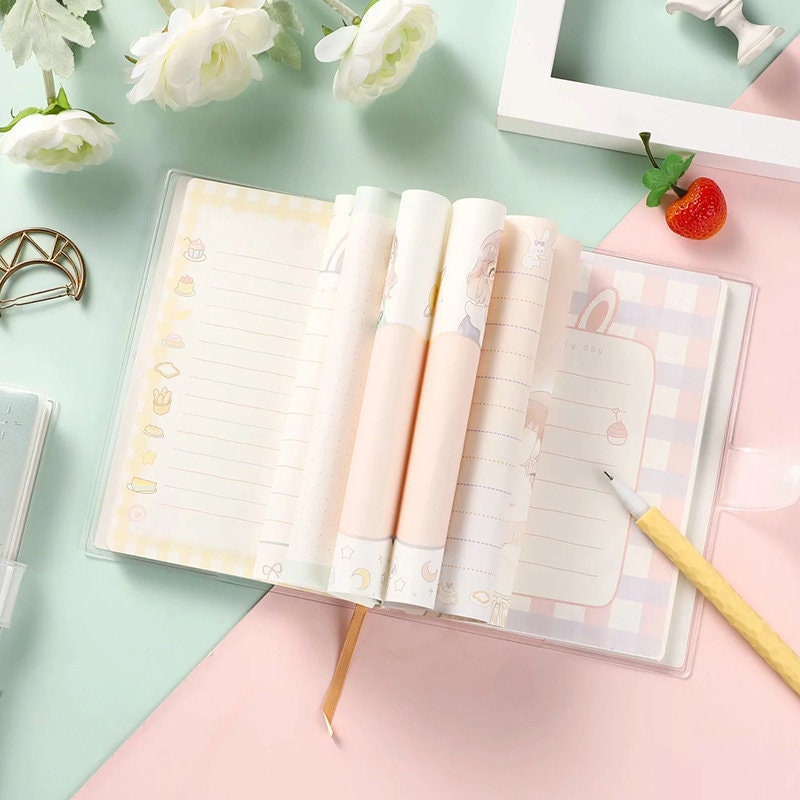 Kawaii Girl PVC Notebook with Buckle and 256 Pages featuring pastel-colored pages, cute illustrations, and decorative elements, displayed with a yellow pencil and floral decor on a pastel background.