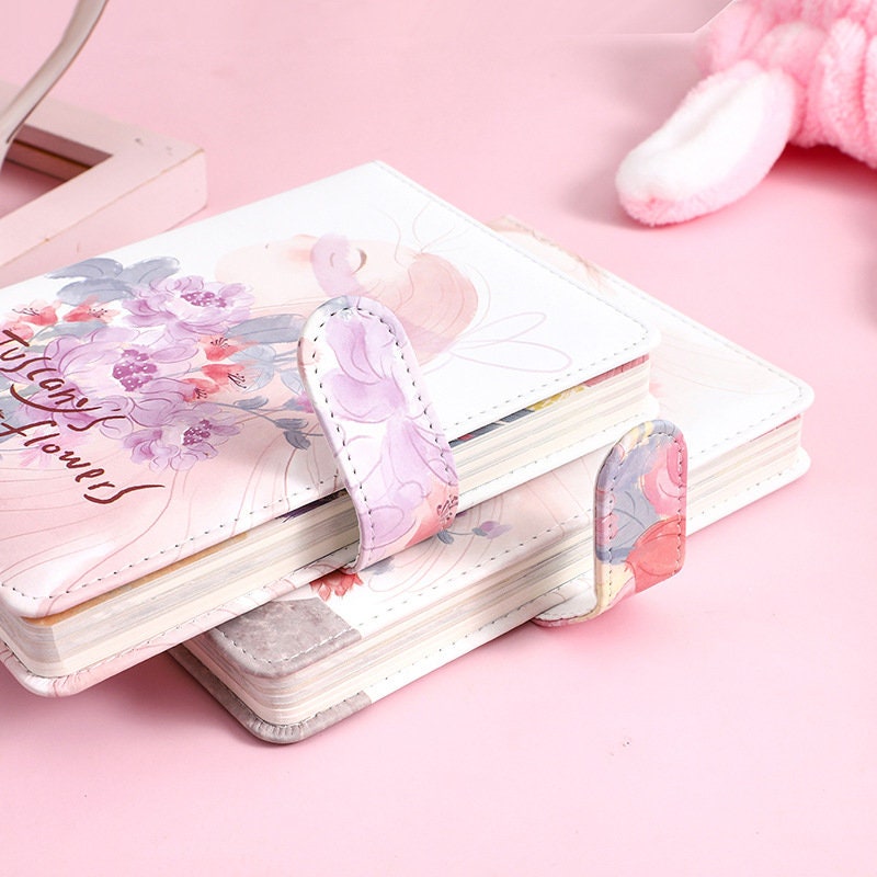 Tuscany's Flowers Leather Journal with Magnetic Buckle, featuring pink floral designs on a white background, displayed on a pastel pink surface.