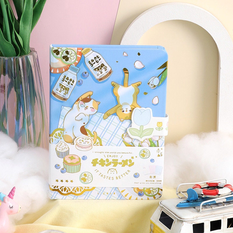 Magnetic Buckle Leather Journal with cute cat illustrations and "It Tastes Better" text, surrounded by colorful kawaii stationery on a pastel background.