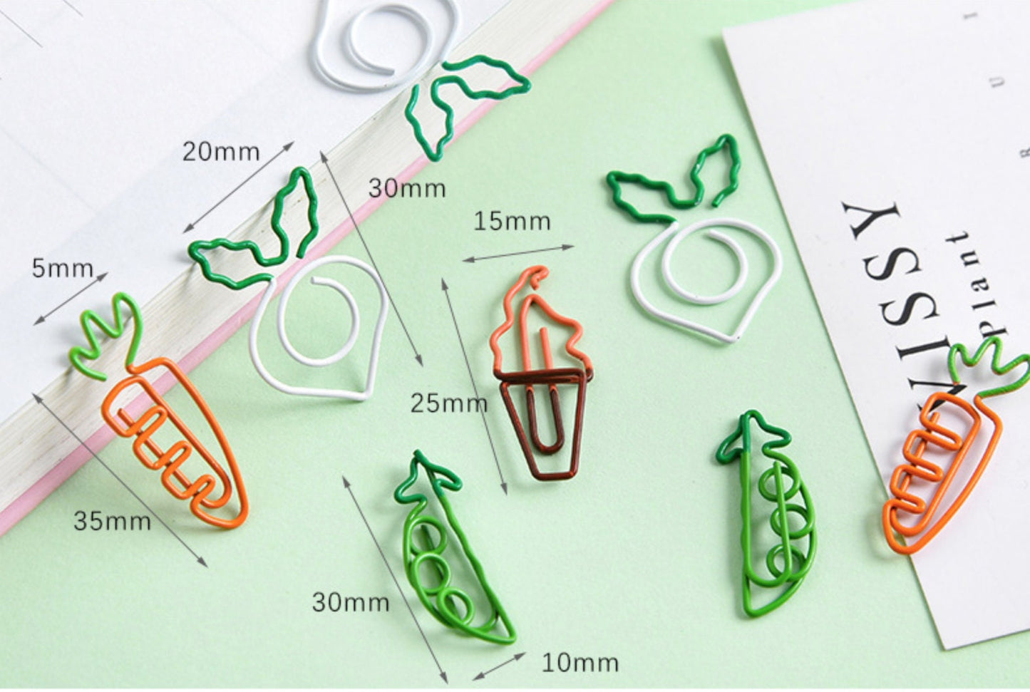 Assorted creative vegetable-shaped clips including carrot, radish, and pea pod designs in green, orange, and white colors, displayed on a pastel background with measurement labels. Cute stationery and office supplies for organizing documents.