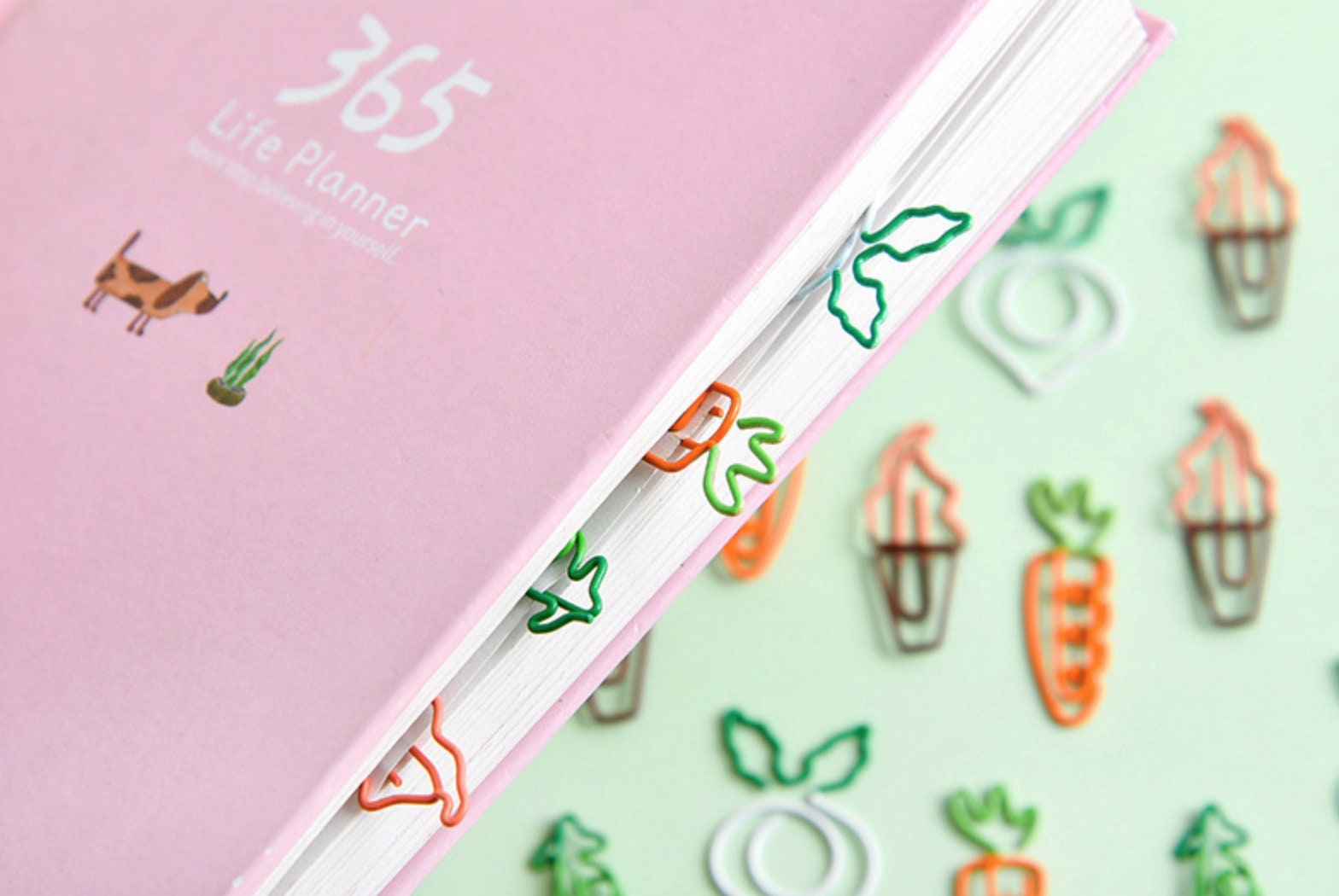 Creative vegetable clips in the shape of carrots and leafy greens used to mark pages in a pastel pink 365 life planner, cute stationery, office supplies.