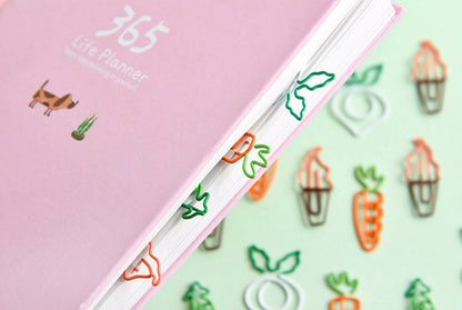 Creative vegetable clips in the shape of carrots and leafy greens used to mark pages in a pastel pink 365 life planner, cute stationery, office supplies.