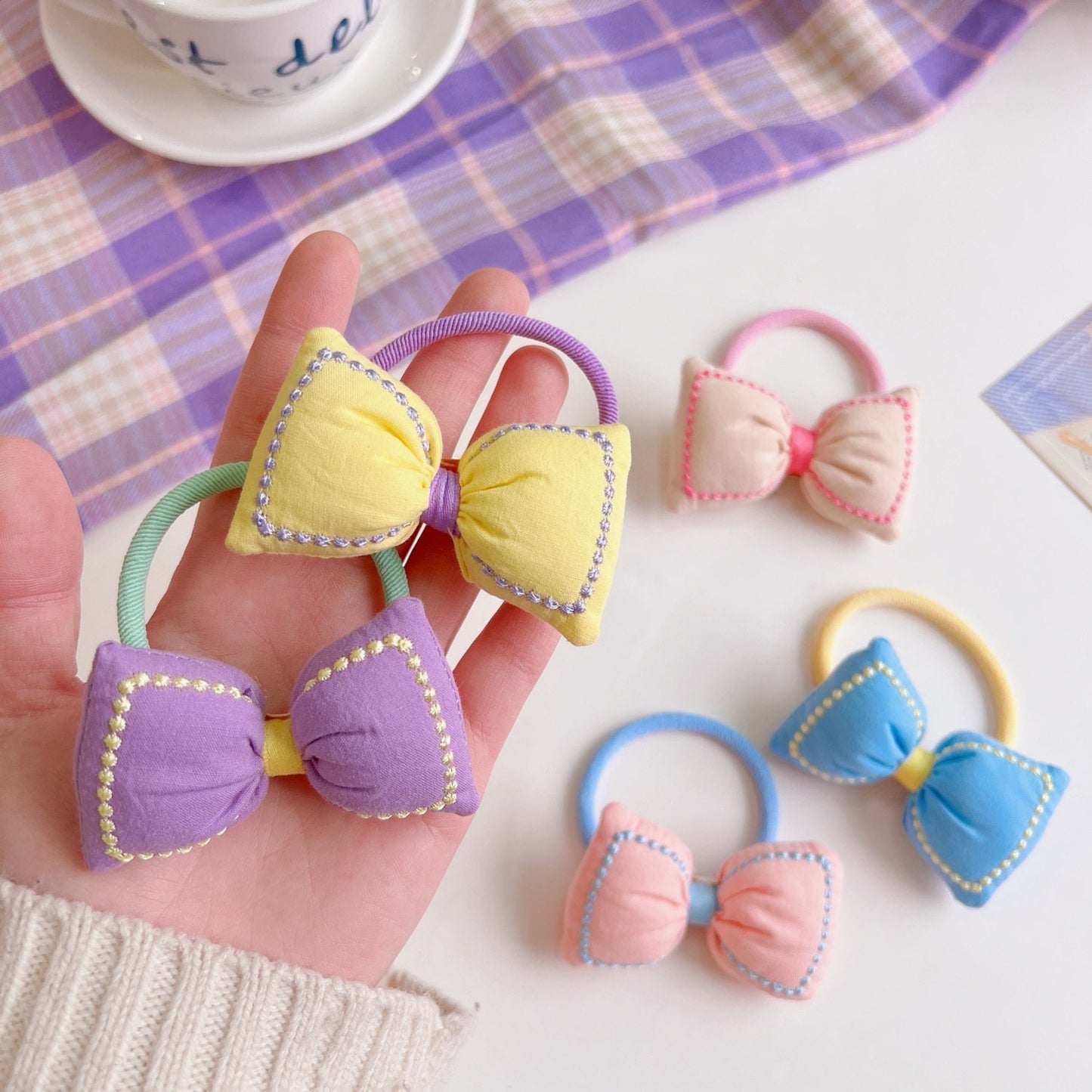 Kawaii bowknot hair tie and bracelet combo in assorted pastel colors including yellow, purple, pink, blue, and beige, displayed on a hand and a table with a plaid cloth backdrop.