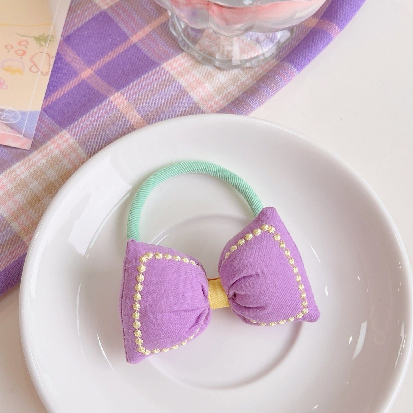 Kawaii Bowknot Hair Tie and Bracelet Combo in lavender and mint green presented on a white plate, featuring cute embroidered edges and a pastel checkered background.