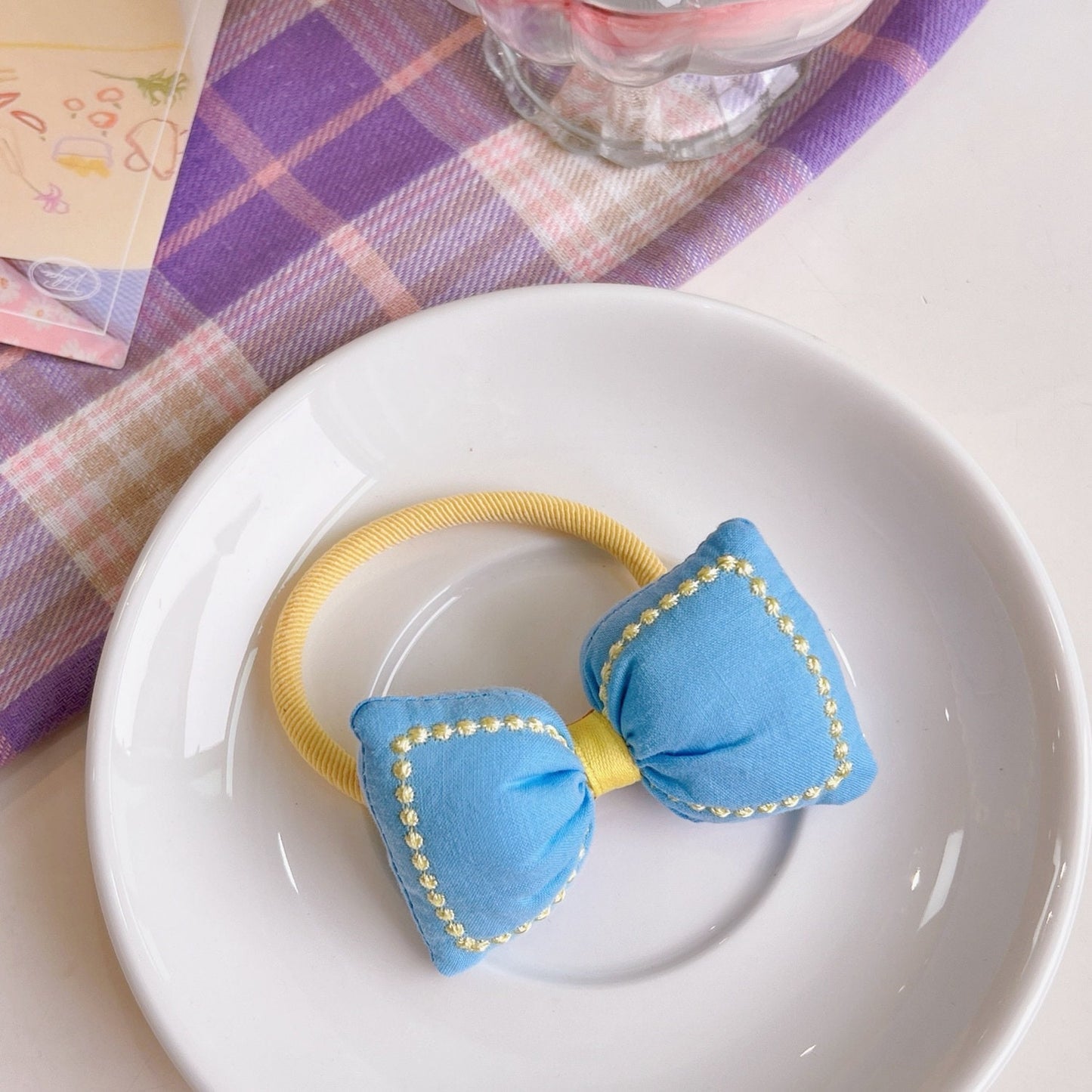 Kawaii Bowknot Hair Tie and Bracelet Combo in blue with yellow elastic band and embroidered detailing, displayed on a white plate with a purple plaid cloth background.