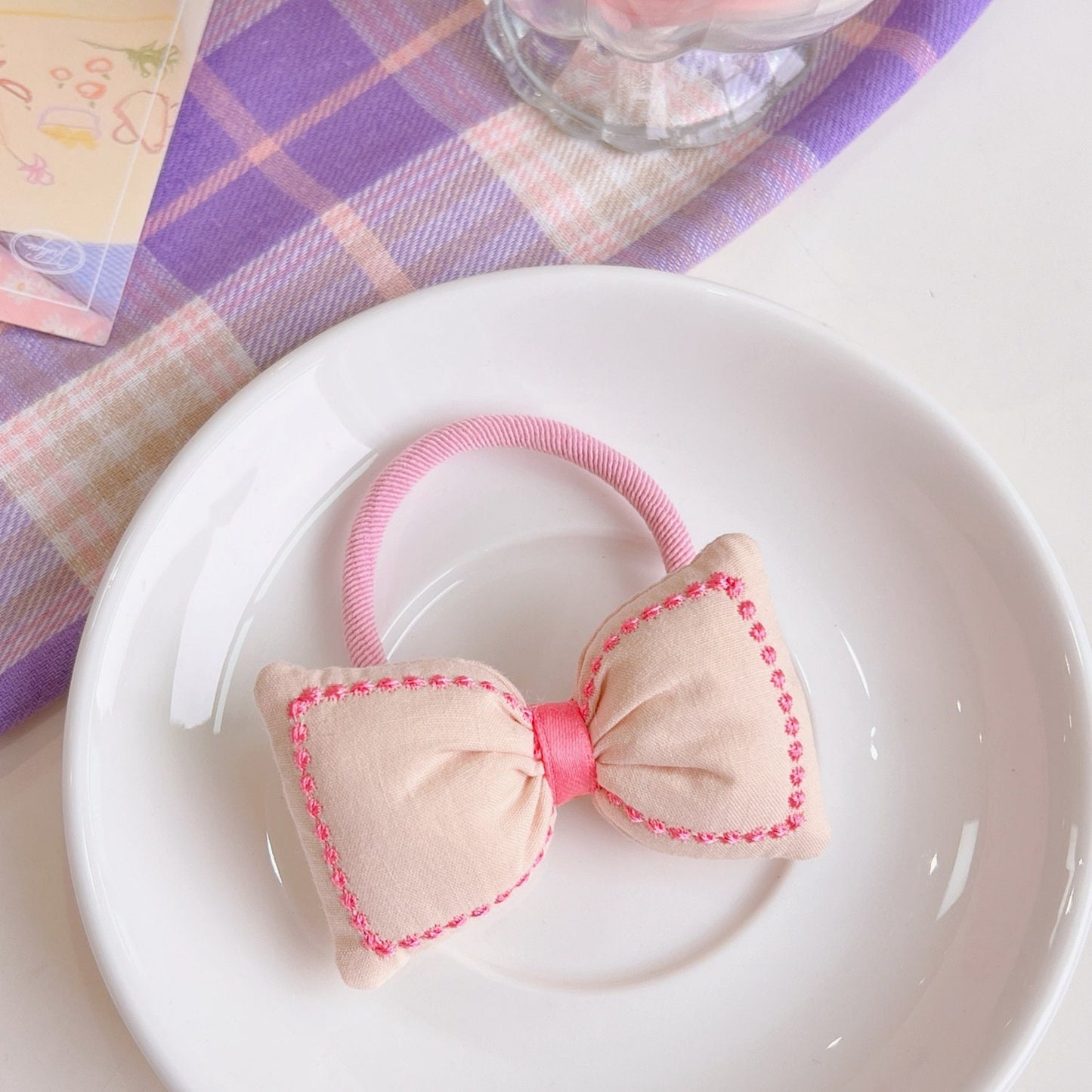 Kawaii Bowknot Hair Tie and Bracelet Combo in pastel pink with delicate stitching detail on a white plate, featuring a plaid purple and pink background.