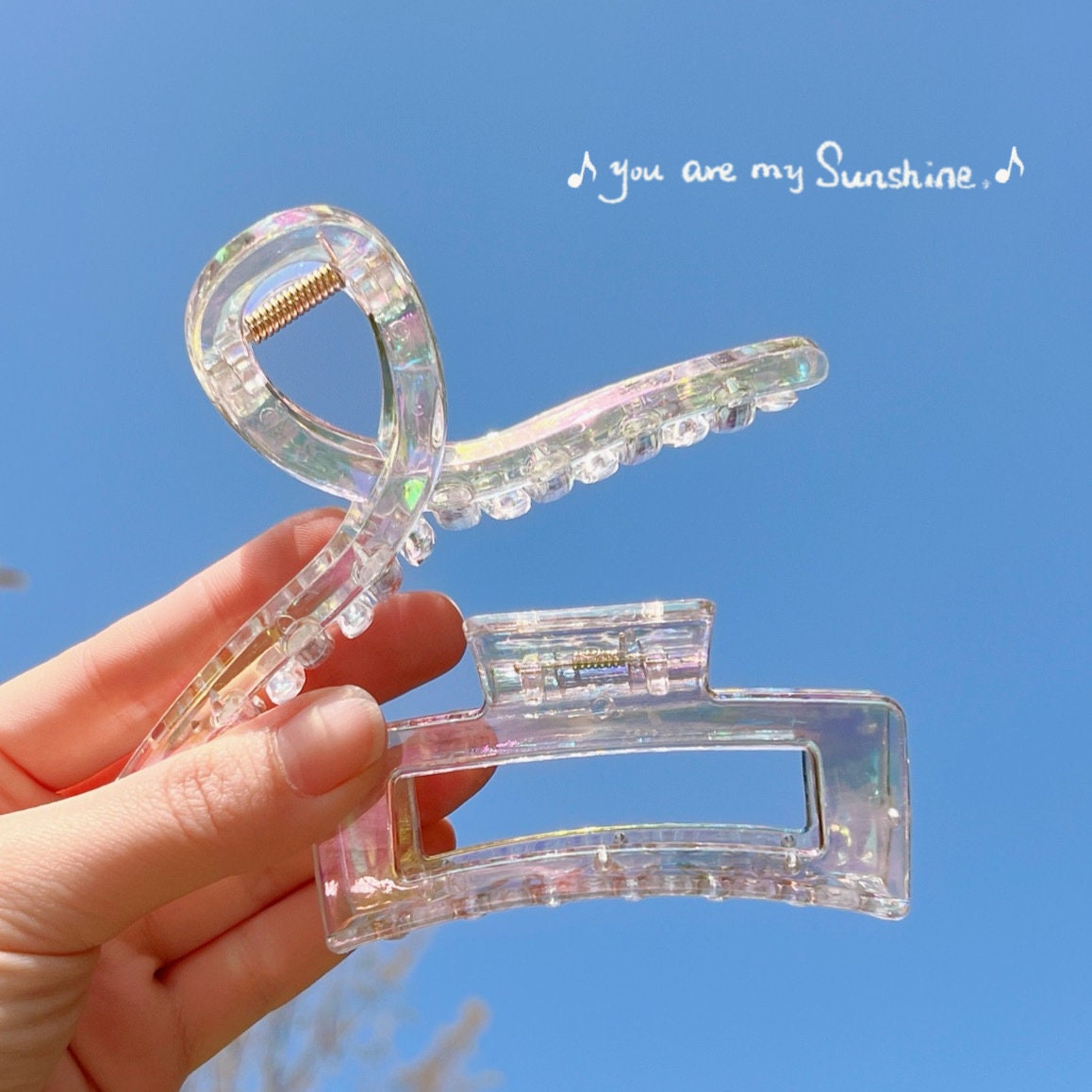 Transparent rainbow acrylic hair claw clip held up against a clear blue sky with the phrase "You are my sunshine" in the background.