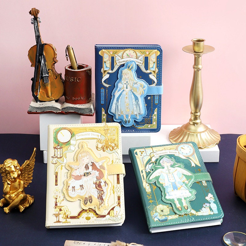 Romantic Dream Leather Journal with Magnetic Buckle A5 Notebook in kawaii style, featuring elegant illustrations and surrounded by decorative items like a violin, candle holder, and angel statue.