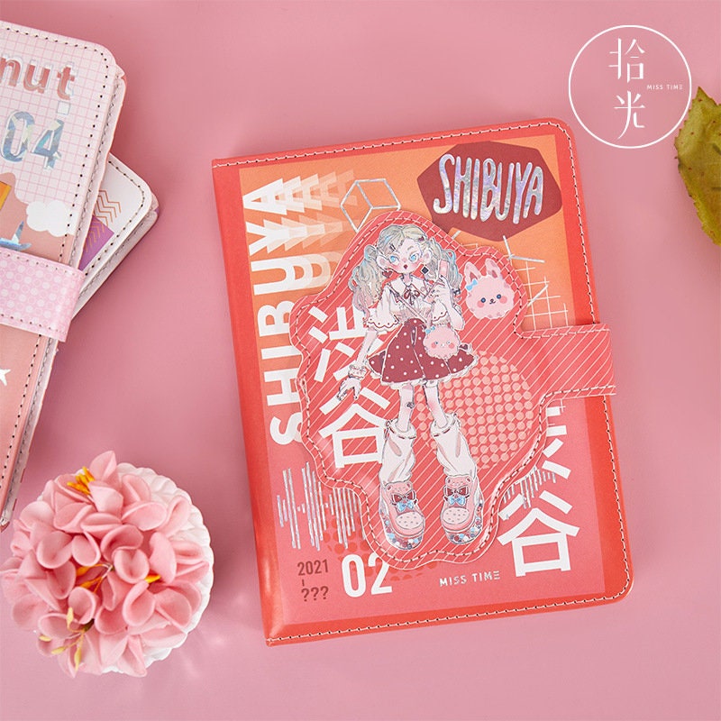 Kawaii Harajuku Leather Journal - A5 Magnetic Buckle featuring a cute anime girl design with "Shibuya" text on a pink and orange background, surrounded by other colorful stationery and a pink flower.