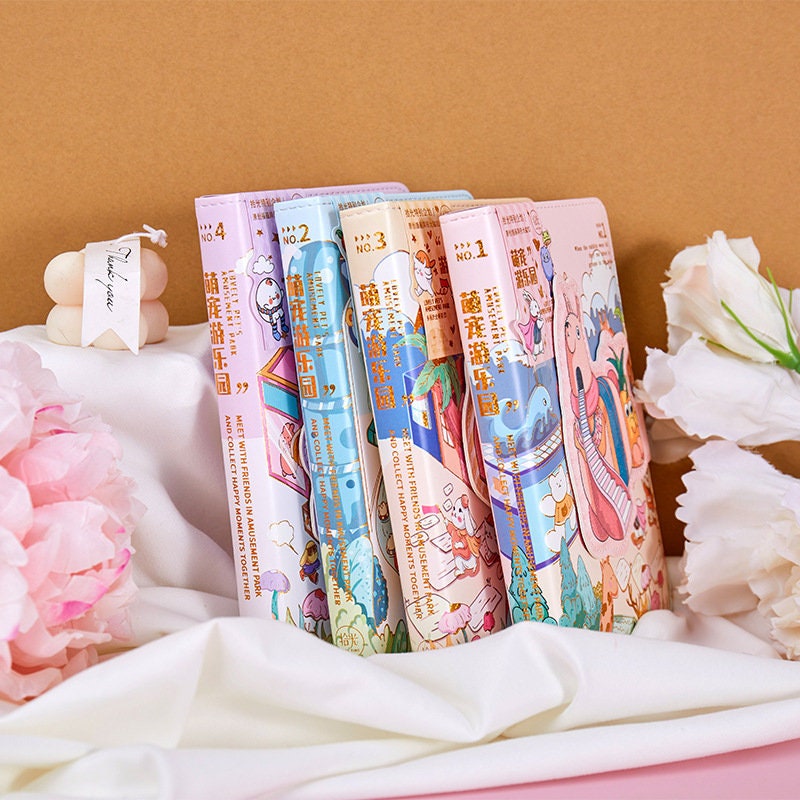 Magnetic Buckle Animal Park Leather Notebooks displayed in various pastel colors with cute animal illustrations, set on a white cloth with pink and white flowers in the background.