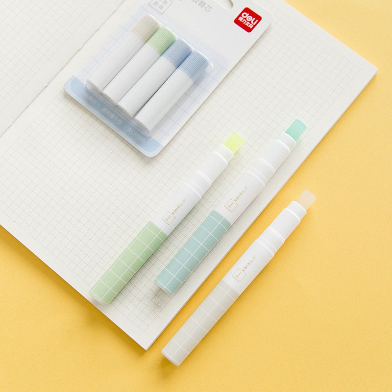 Kawaii glue stick for scrapbooking and journaling, featuring pastel-colored caps and grid design, displayed on a gridded notebook with a pack of three glue sticks and individual sticks.