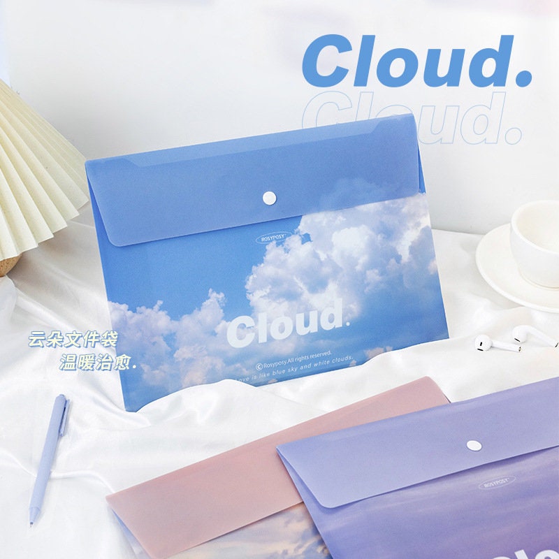 Portable A4 file folder with button closure, featuring a sky and clouds design, displayed on a desk with other stationary items.