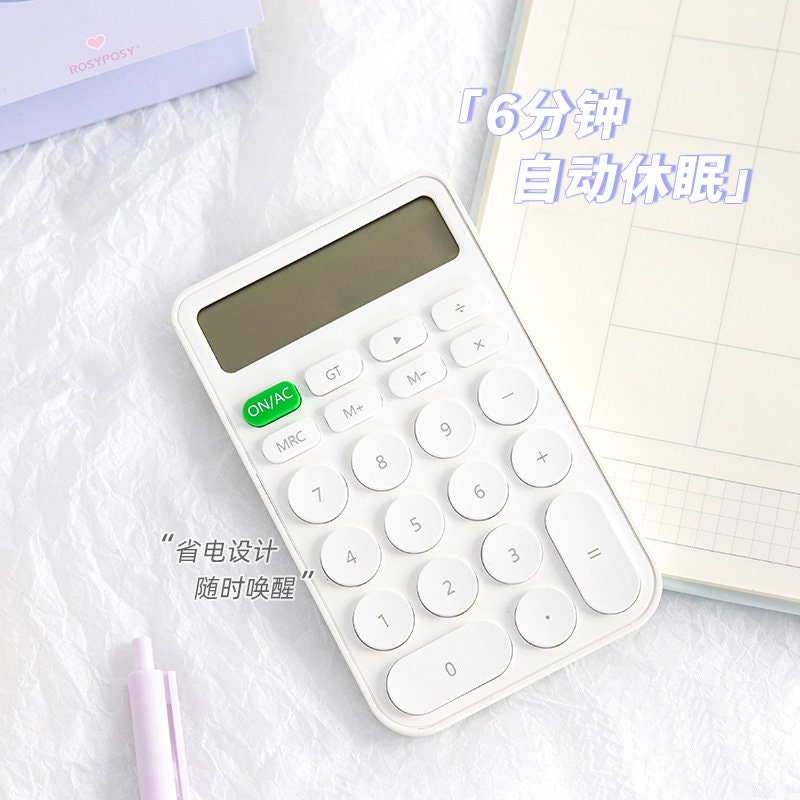 Big Button Cute Electronic Calculator in white with a large display, green ON/AC button, kawaii design, resting on a desk next to a grid notebook and a pink pen.