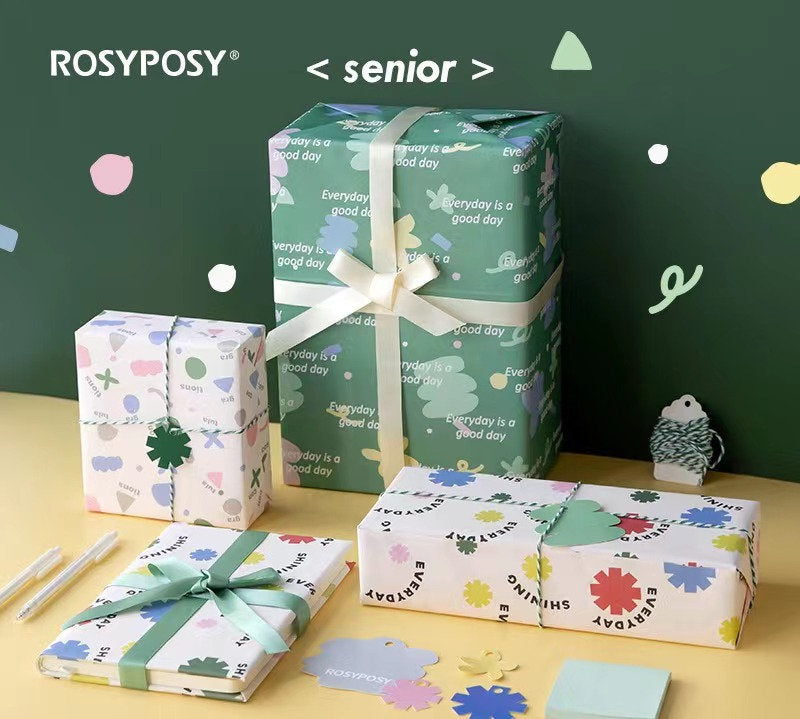 Colorful gift boxes wrapped with vibrant patterned gift wrapping paper featuring various designs including "Everyday is a good day," floral patterns, and pastel shapes, adorned with ribbons and bows, surrounded by pens and paper cutouts on a yellow surface.